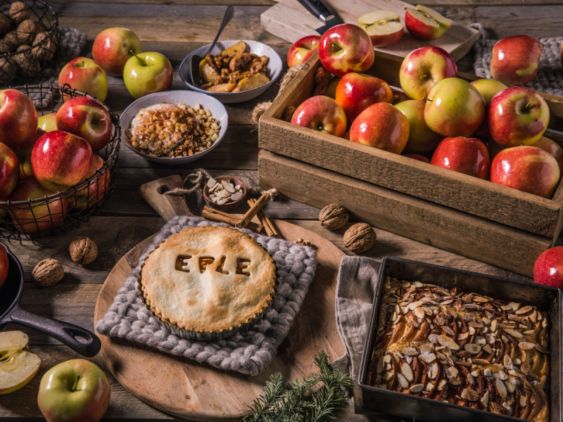 Eplepai med epler og høstlige dekorasjoner på et bord, omgitt av friske epler og nøtter.