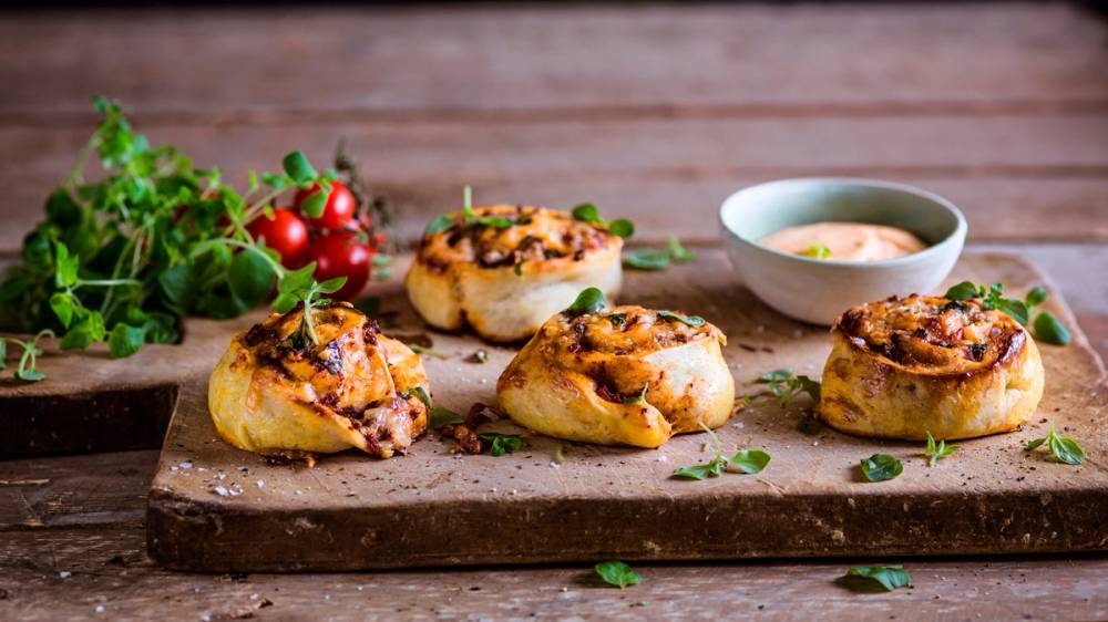 Fire kjøttdeigpizzasnurrer på en skjærefjøl med en liten skål saus ved siden av, dekorert med frisk urter og tomater i bakgrunnen.