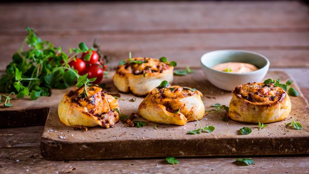 Fire kjøttdeigpizzasnurrer på en skjærefjøl med en liten skål saus ved siden av, dekorert med frisk urter og tomater i bakgrunnen.