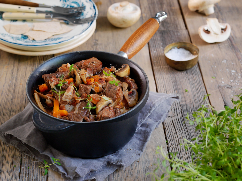 En gryte med biff bourguignon med kjøtt, gulrøtter og sopp, plassert på et bord med en skje og noen krydderurter ved siden av.