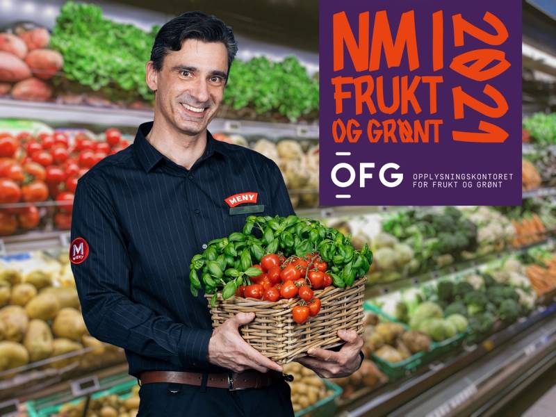 Person i butikkholder en kurv med tomater og grønnsaker foran en frukt- og grønnsaksdisk, med en plakat som sier 'NM i frukt og grønt 2023'.