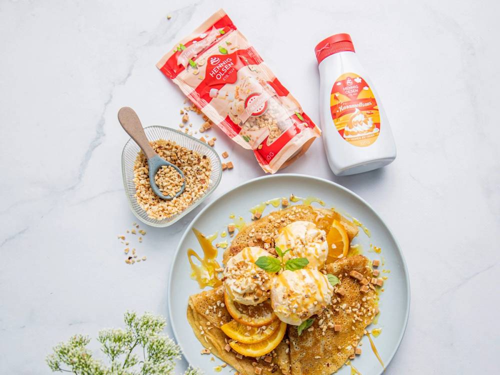 En tallerken med iskrem og bananer, et glass med saus, og en pose med snacks på et hvitt bord.