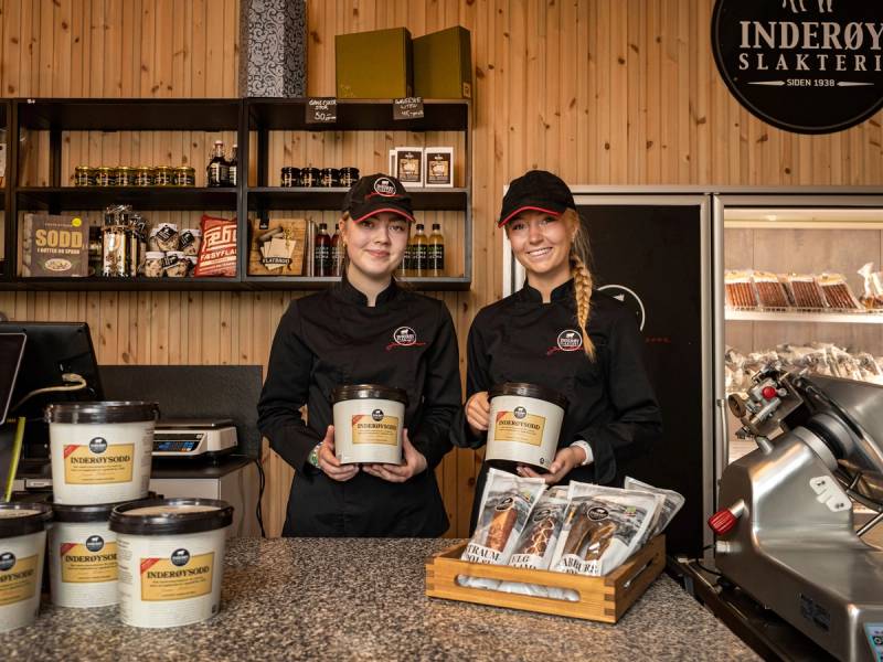 To ansatte i slakteriet står bak disken med produkter og en kjøttmaskin, med et skilt for Inderøy Slakteri på veggen bak dem.