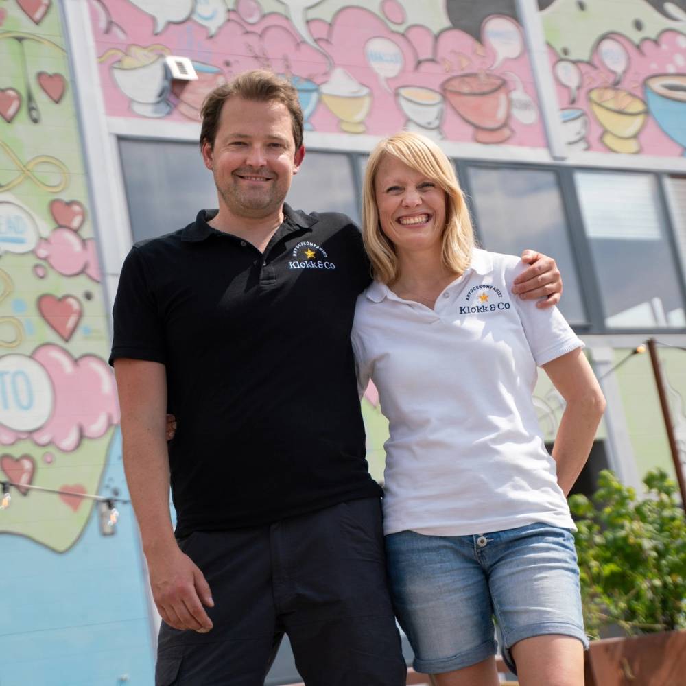 To personer står sammen foran en fargerik vegg med hjerter og kaker, og de har armene rundt hverandre.