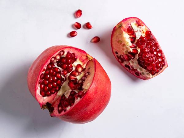 Et halvt granateple med mange røde frø og noen løse frø rundt på en hvit bakgrunn.