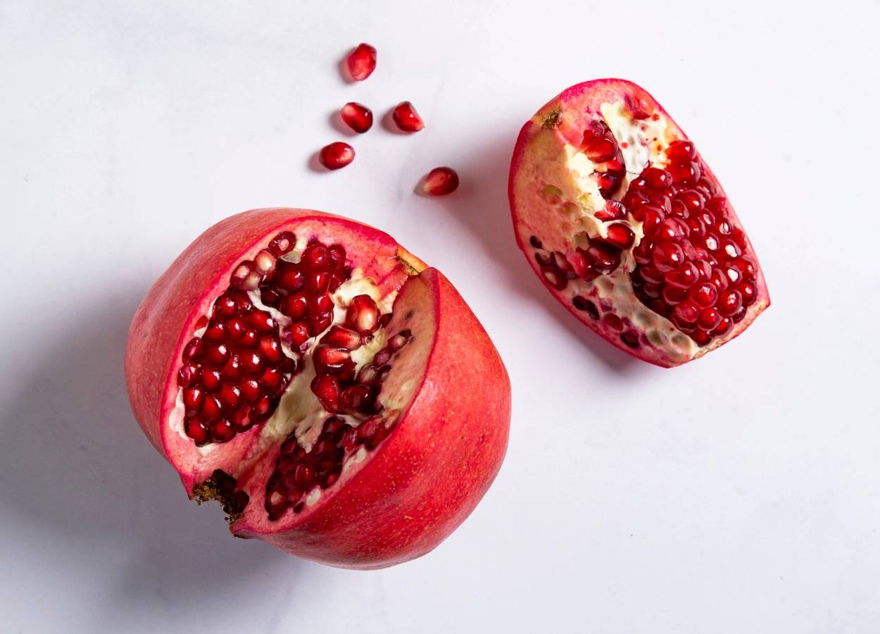 Et halvt granateple med mange røde frø og noen løse frø rundt på en hvit bakgrunn.
