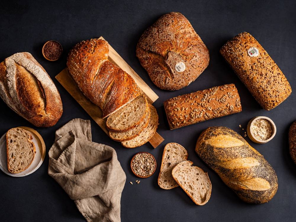 Ulike typer brød og baguetter ligger på et mørkt bord med en liten skål med smør og en klut ved siden av.