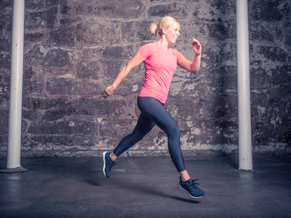 En kvinne hopper i en treningshall med en betongvegg i bakgrunnen, iført rosa treningstøy og svarte treningssko.