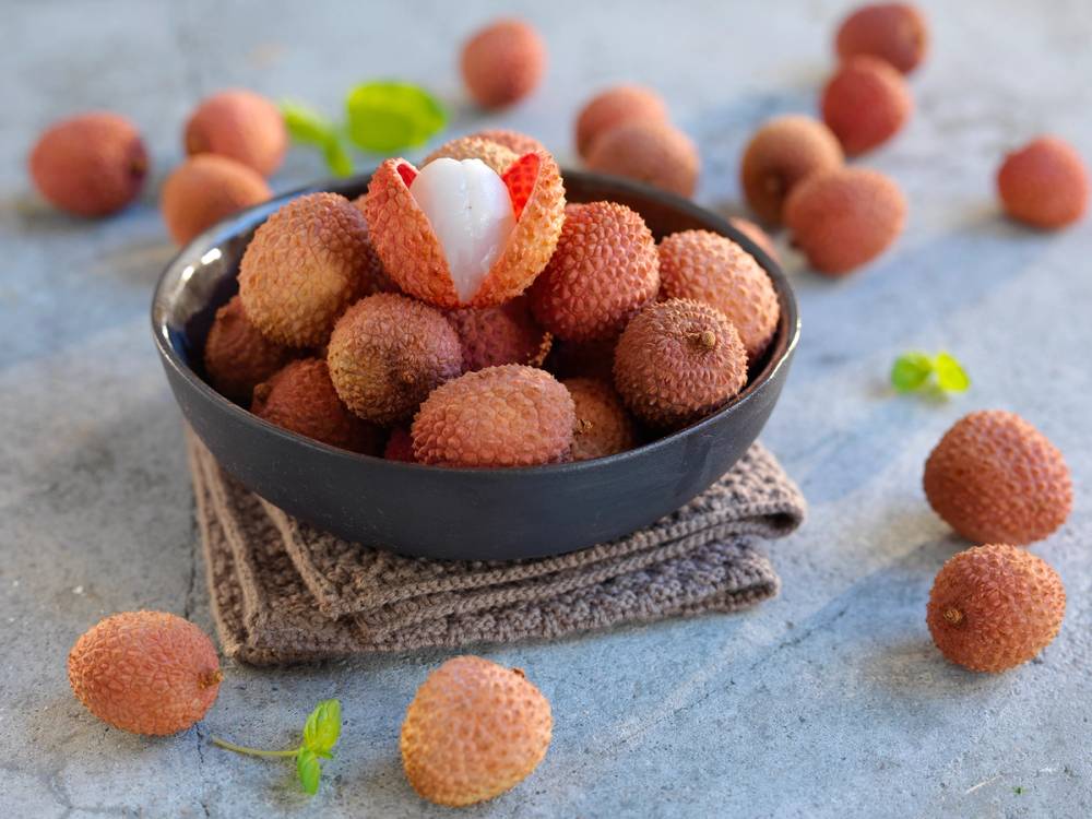 En skål med lychee-frukter, noen av dem skrellet og oppdelt, på et bord med noen spredte frukter rundt.