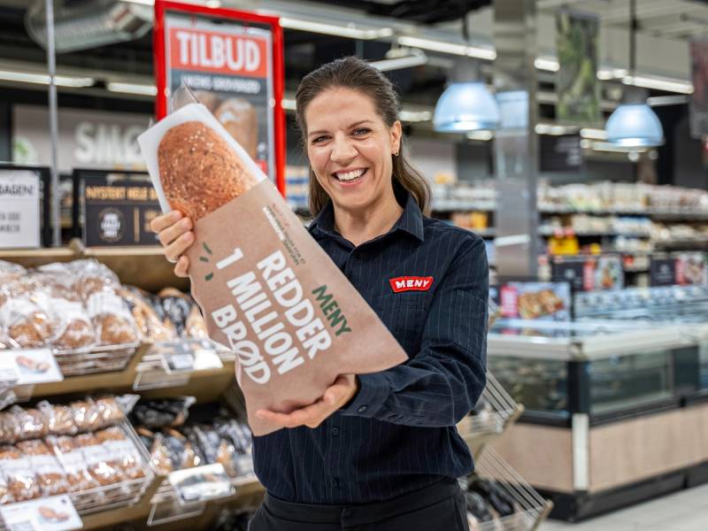 Kvinne i butikk med en pose med brød, med hyller og tilbudsskilt i bakgrunnen.