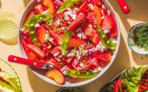 En fargerik sommersalat med jordbær, tomater og grønne chilipepper i en bolle, omgitt av salat og kjøkkenutstyr.