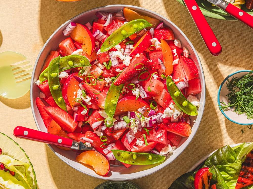 En fargerik sommersalat med jordbær, tomater og grønne chilipepper i en bolle, omgitt av salat og kjøkkenutstyr.