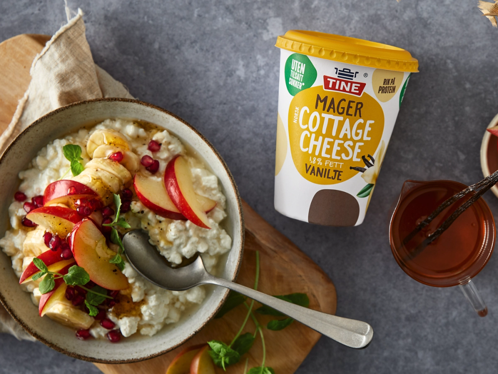 En skål med cottage cheese toppet med vanilje, frukt og bær, sammen med en kopp te eller kaffe, og en pakke med vanilje-cottage cheese ved siden av.