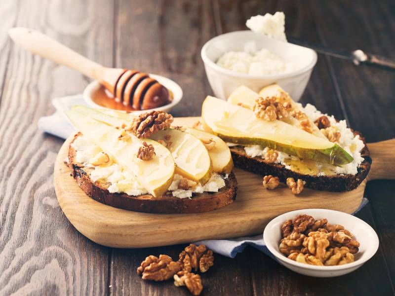 Bruschetta med pære, ricottaost og valnøtter på et trebord med honning og ekstra valnøtter ved siden av.