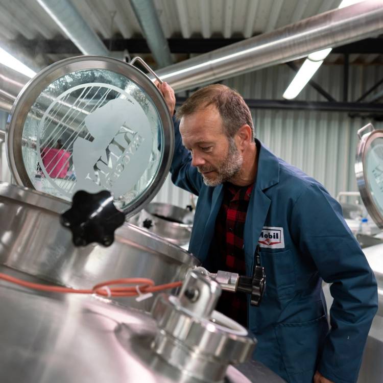 En person med skjult ansikt undersøker en stor metalltank i et industrielt miljø, sannsynligvis i en matproduksjonsfabrikk.