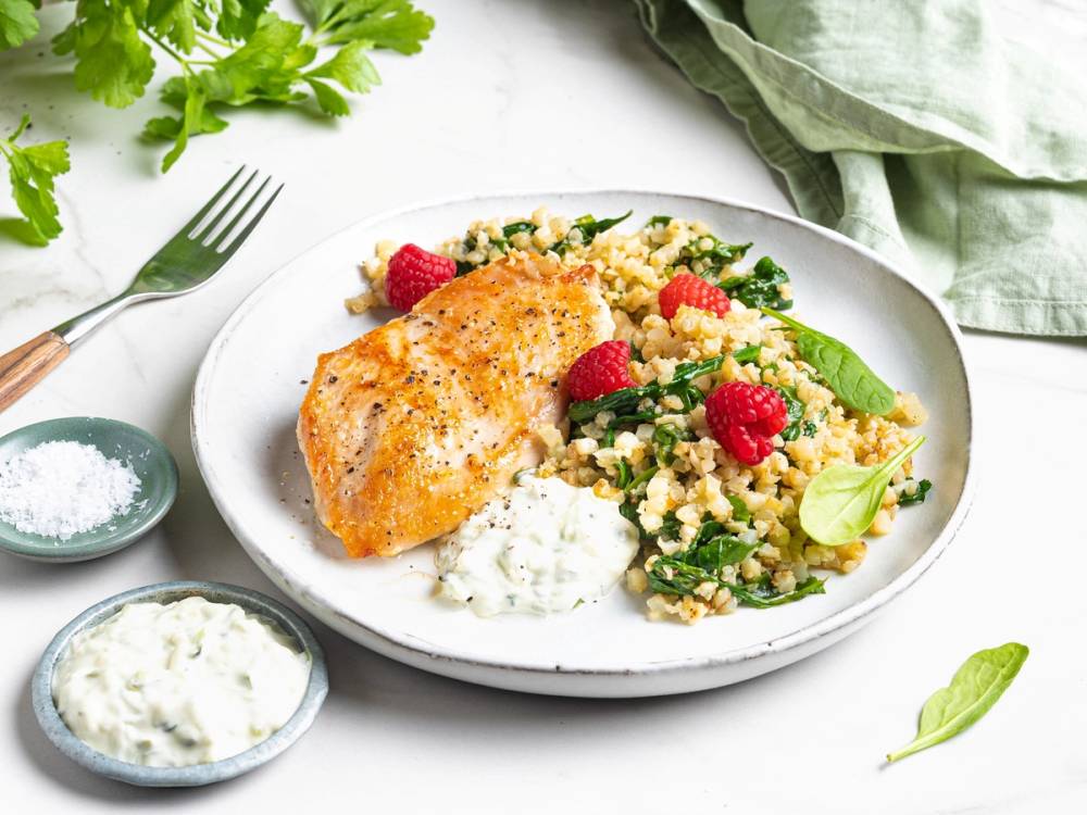 Kyllingfilet med blomkålris og friske bringebær på en hvit tallerken, omgitt av urter og små skåler med saus og krydder.