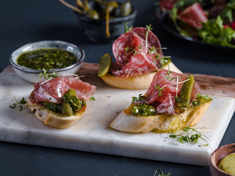 To små bruscettabrød med skinke, pesto og små pickles på en skjærebrett, omgitt av salat og en skål med pesto i bakgrunnen.