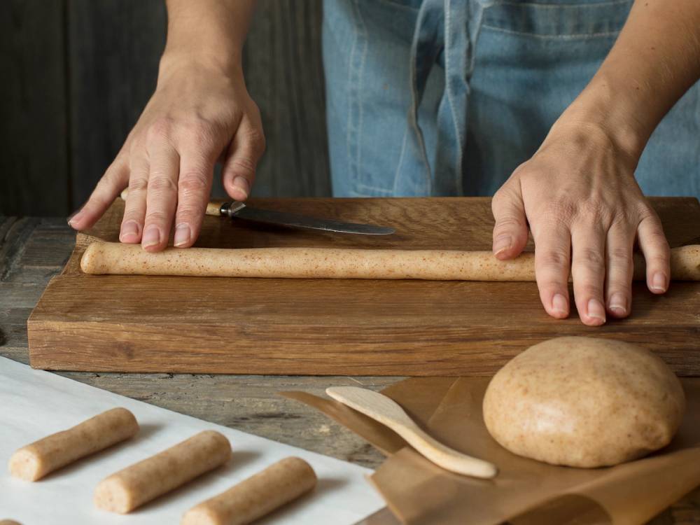 Person skjærer en kransekakestang med en kniv på et kjøkkenbord, omgitt av små kaker og en bolle med deig.