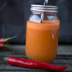 Et glass med oransje juice eller smoothie med sugerør, plassert på en treflate med en rød chili ved siden av.