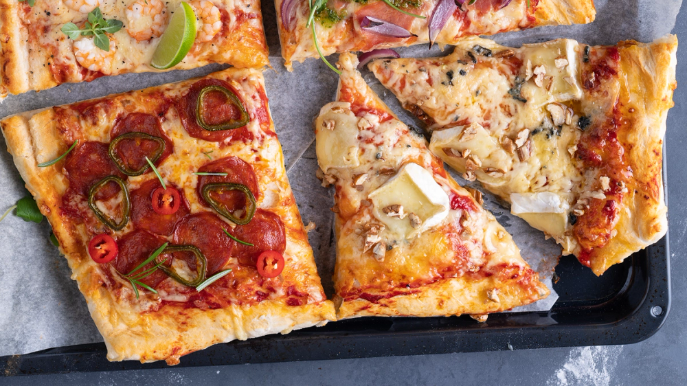 To stykker pizza på en langpanne, delt i fire, med ulike toppings som pepperoni, grønnsaker og ost, på et svart bakepapir.