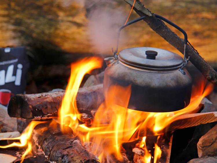 En svart kjele som kokes over åpen ild med flammer og røykskyer, omgitt av steiner og trestykker.