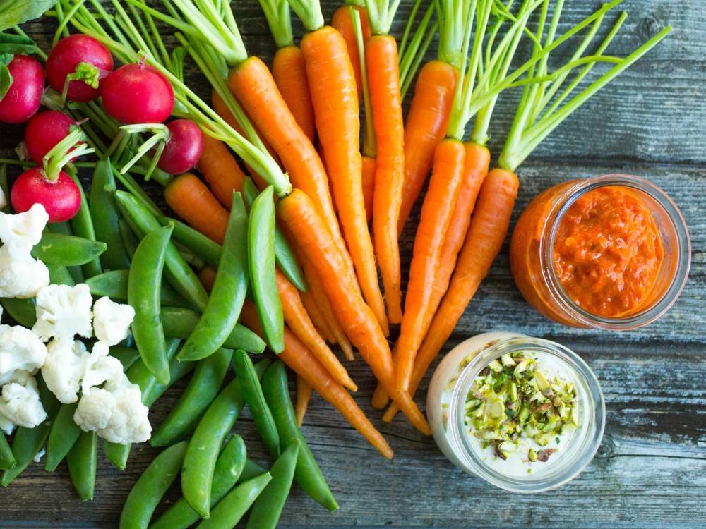 Fargerike grønnsaker som gulrøtter, reddiker, sukkererter og blomkål med to små glass med dip, på en treplate.