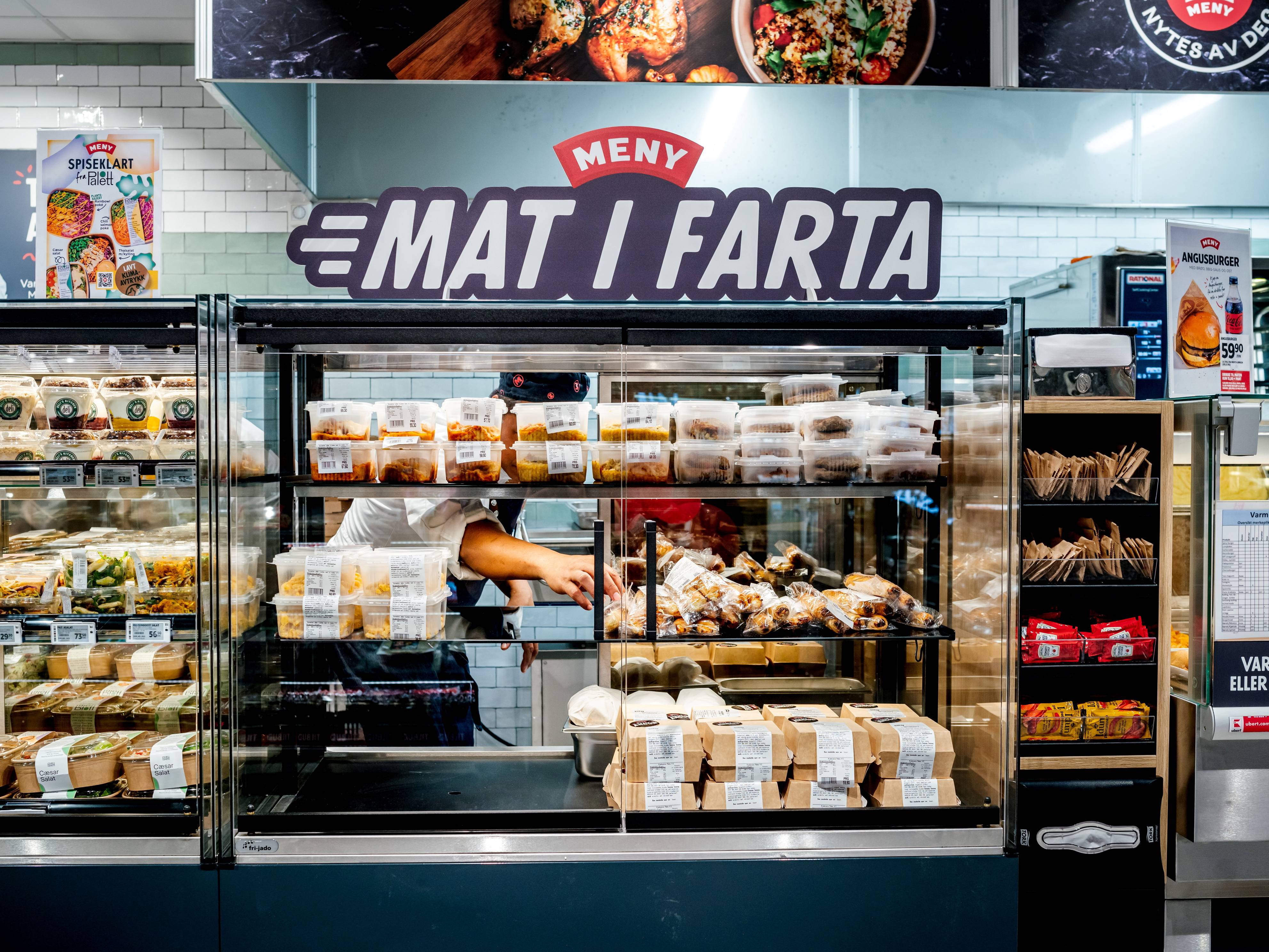 En matbutikk med en kjøledisk som selger brød og bakervarer, og et skilt som sier 'Meny' og 'Mat i fart' i bakgrunnen.