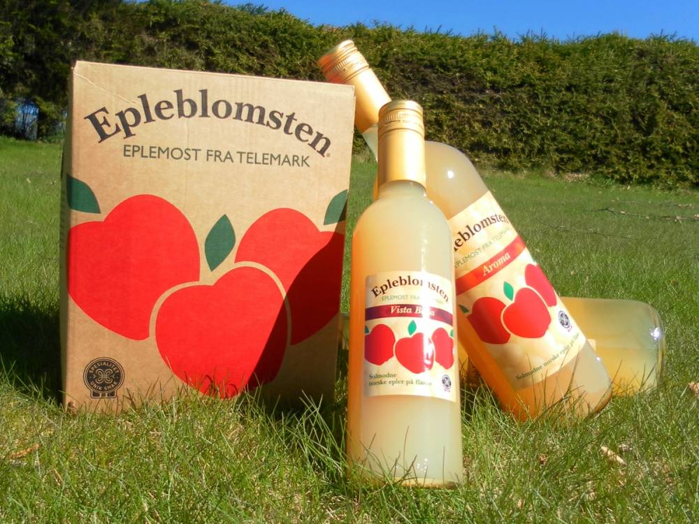 To flasker og en eske med epleblomstdrikk på gress med busker i bakgrunnen