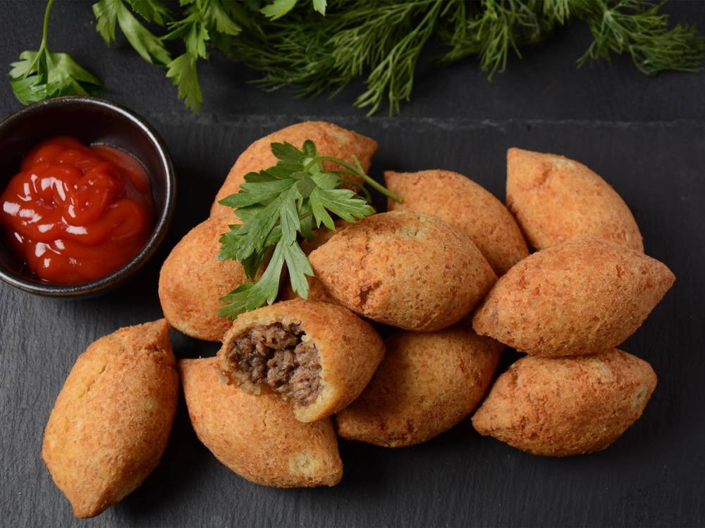 Kibbeh, små ovnsbakte kuler med kjøttfyll, servert med en skål med tomatsaus og fersk grønn urt på en svart bakgrunn.