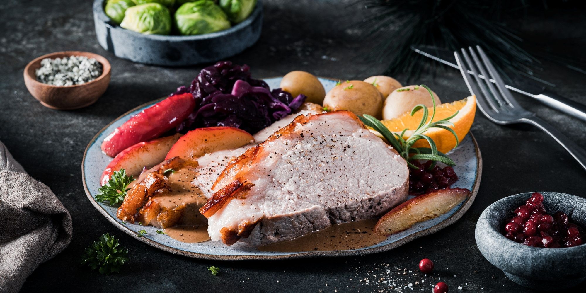 Skinkestek servert på tallerken med poteter, rosenkål og tyttebær, omgitt av grønnsaker og krydderurter.