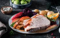 Skinkestek servert på tallerken med poteter, rosenkål og tyttebær, omgitt av grønnsaker og krydderurter.