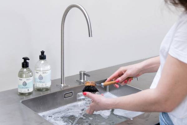 En person vasker en svart gjenstand i en kjøkkenvask med vann og såpe, mens to såpeflasker står på benken ved siden av.