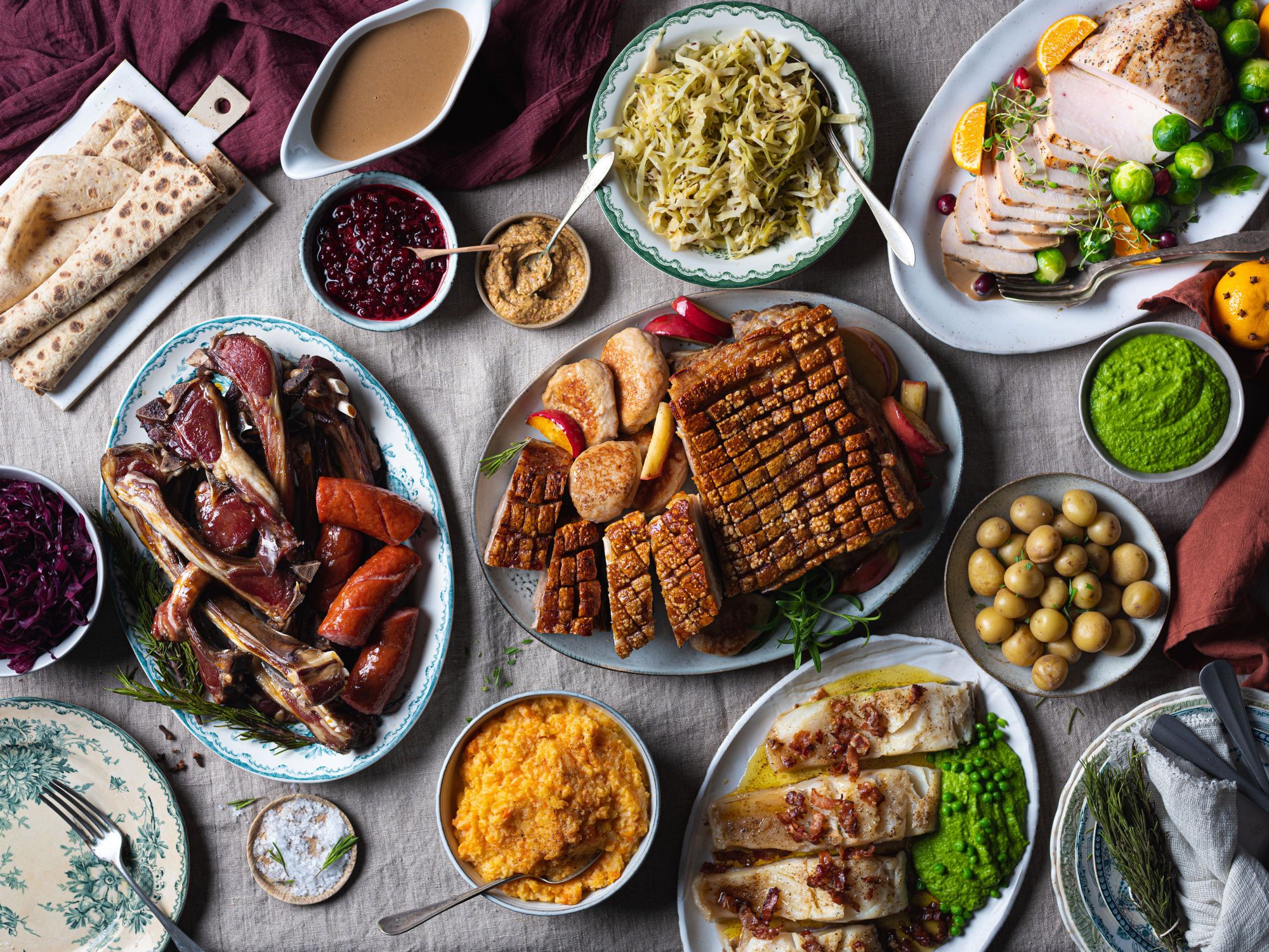 Et utvalg av tradisjonell julemat, inkludert ribbe, medisterkaker, surkål, grønnsaker og julepølser, på et dekket bord.