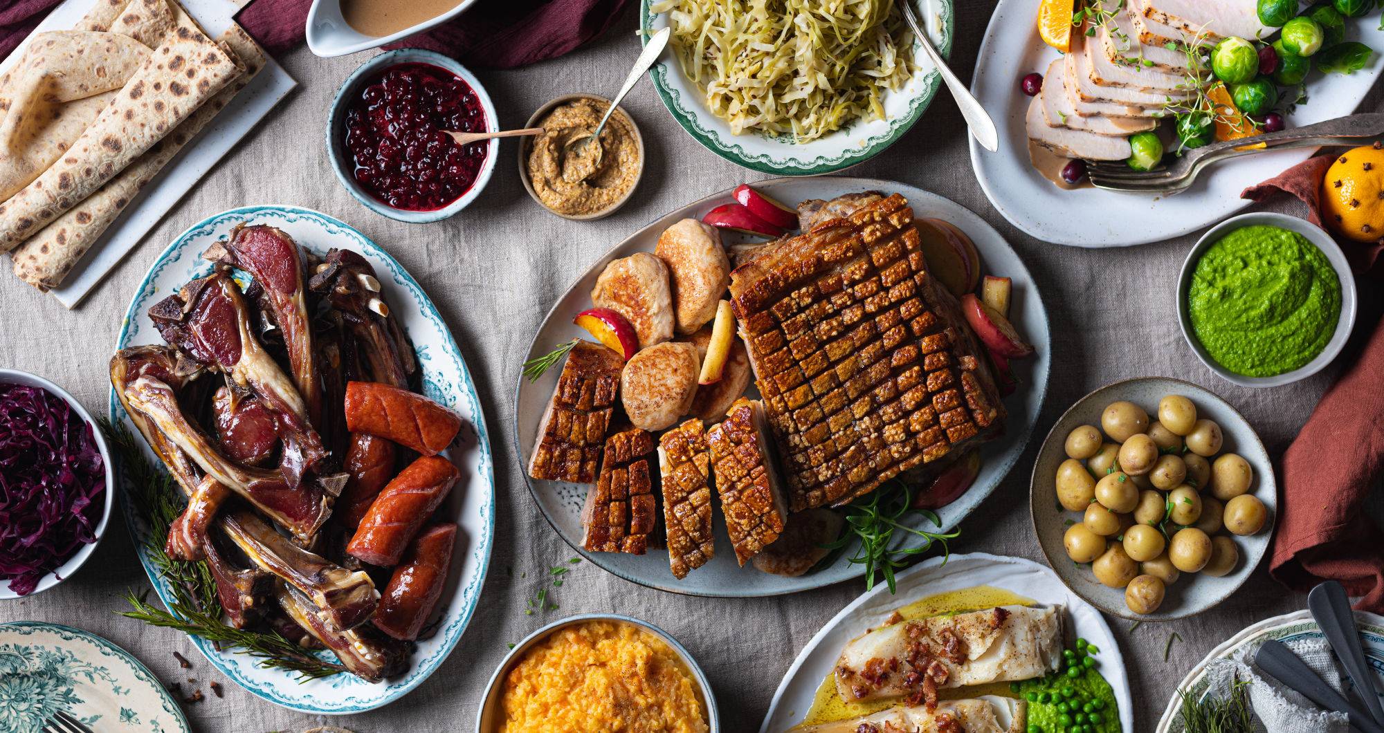 Et utvalg av tradisjonell julemat, inkludert ribbe, medisterkaker, surkål, grønnsaker og julepølser, på et dekket bord.