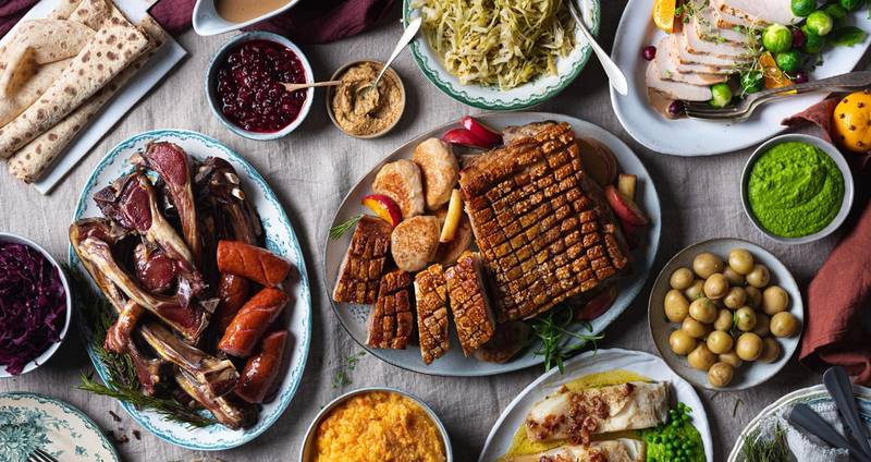 Et utvalg av tradisjonell julemat, inkludert ribbe, medisterkaker, surkål, grønnsaker og julepølser, på et dekket bord.