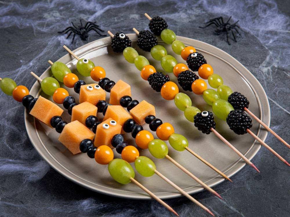 Fargerike fruktspyd med gule, grønne, svarte og oransje biter, pyntet med små svarte edderkopper, på en sølvfarget tallerken til Halloween.