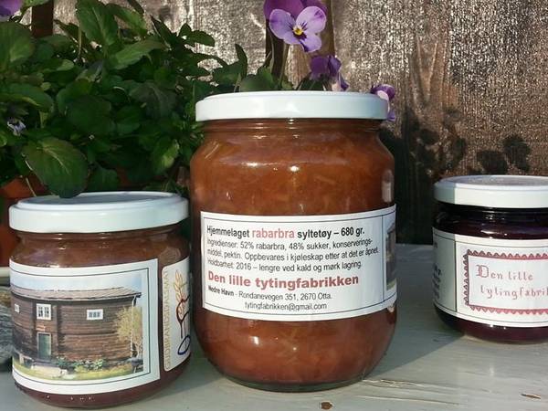 Rabarbrasyltetøy i glasskrukker med etiketter, plassert på en hylle med blomster i bakgrunnen.