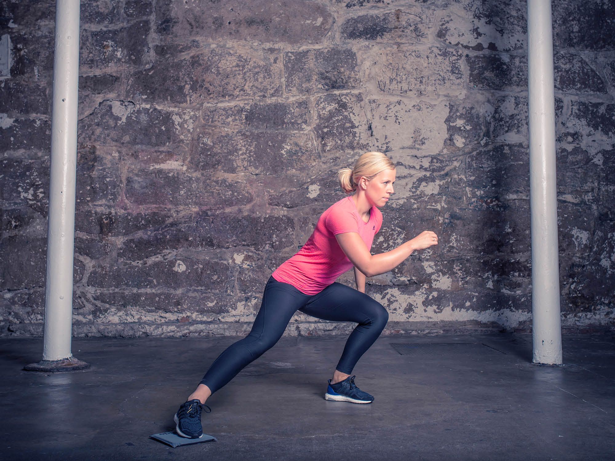 En person i rosa treningtøy gjør utfall foran en murvegg med sklimatte, klar for trening eller øvelse.