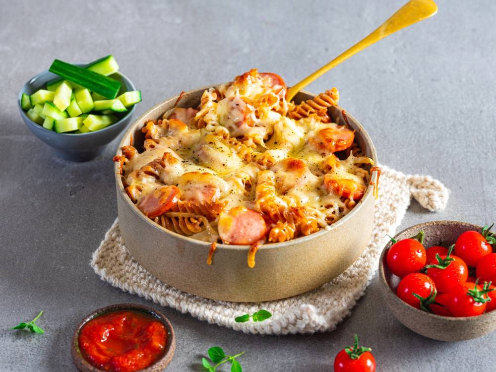 Pølsegryte med ost og grønnsaker i en rund form, servert med en skål med cherrytomater og en liten skål med hakkede grønnsaker ved siden av.