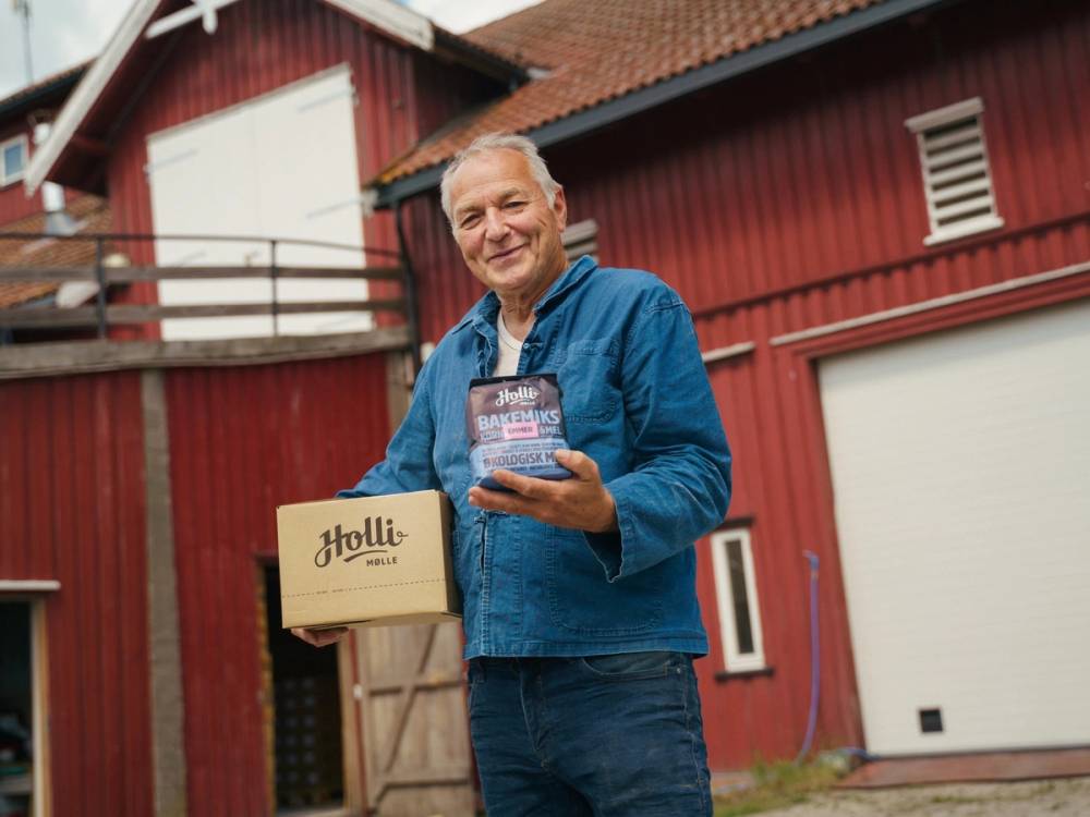 Person holder en pakke med Holli Mølle-brød foran en rød trebygning.