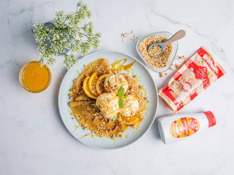 HennigOlsen Snakkis galette med appelsinsaus og karamell servert med et glass appelsinjuice og en liten skål med karamellsaus på et hvitt bord.