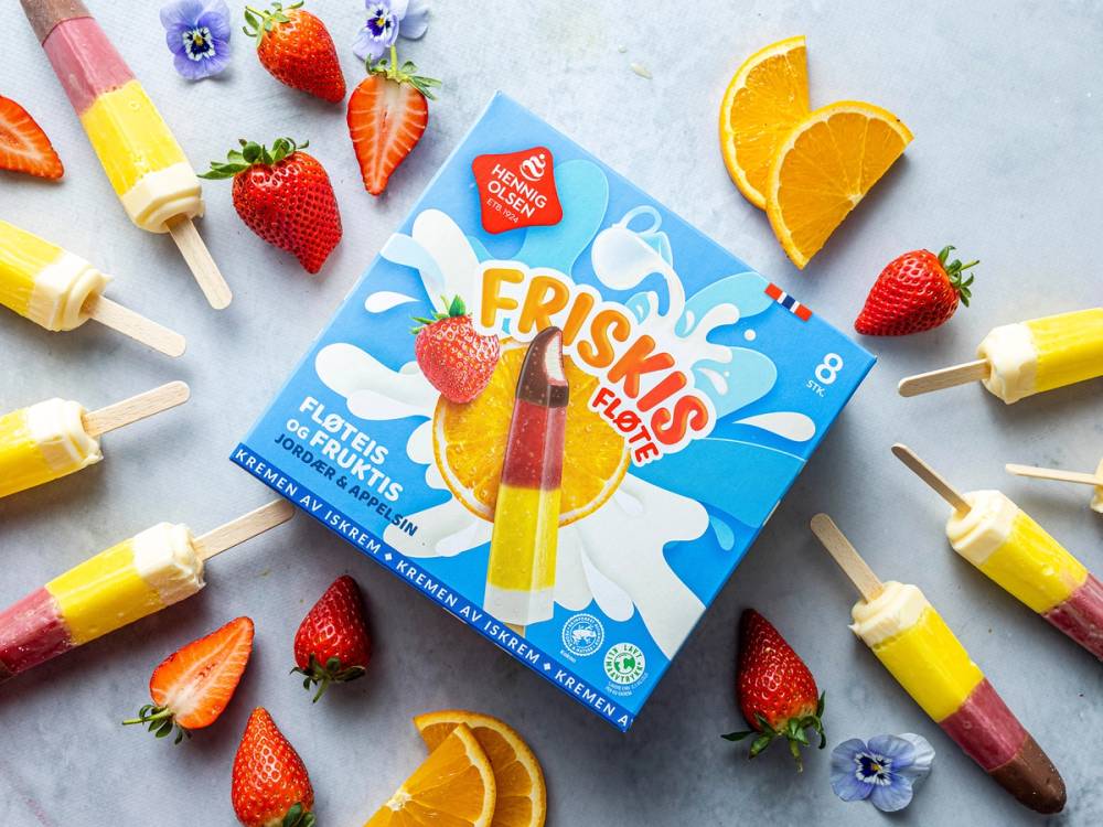 HennigOlsen Friskis iskrem med jordbær og appelsin, omgitt av flere jordbær, appelsinskiver og iskrempinner på et bord.