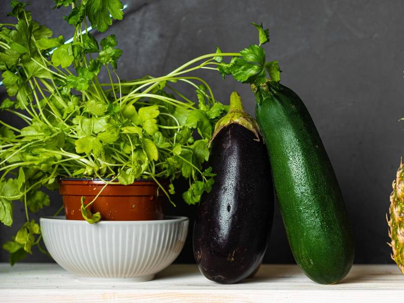 Friske urter i en hvit bolle og to squash, en lilla og en grønn, på et bord.