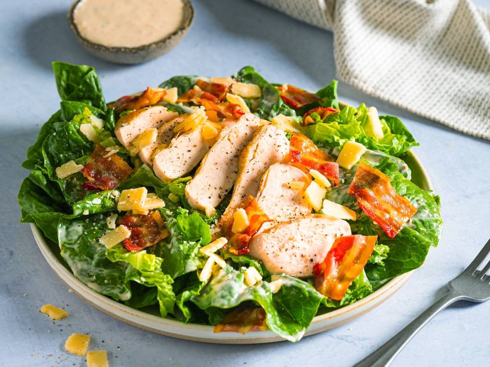 En frisk cesarsalat med kylling, romanosalat, baconbiter og revet ost på en hvit tallerken.
