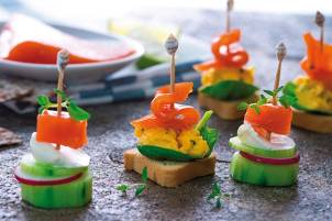 Små kanapeer med røkelaks, kremost, agurk og paprika på små brødskiver, pyntet med små flagg, på et fat.