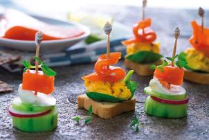 Små kanapeer med røkelaks, kremost, agurk og paprika på små brødskiver, pyntet med små flagg, på et fat.