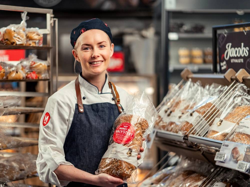 En person i bakeri med hvit skjorte og blå forkle som holder et brød i en butikk. Sikkerhet 90.
