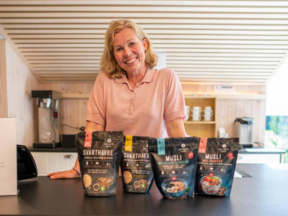 Person står bak en kjøkkenbenk med fire poser med frokostblanding foran seg, i et lyst kjøkken med trepanel på veggen.