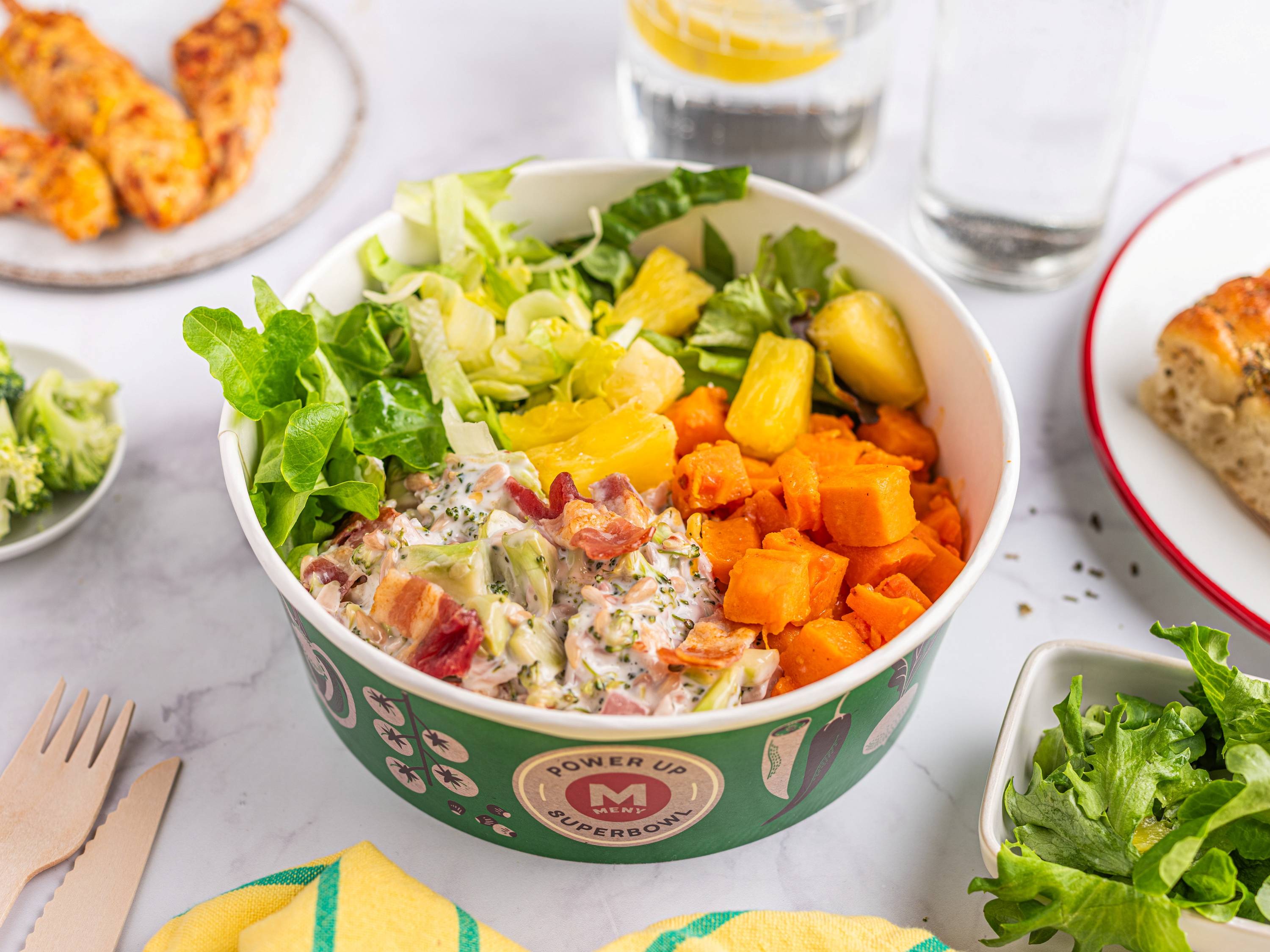 En bolle med salat og frukt, inkludert grønne blader, ananas og gulrot, på et bord med andre retter og drikkevarer rundt.