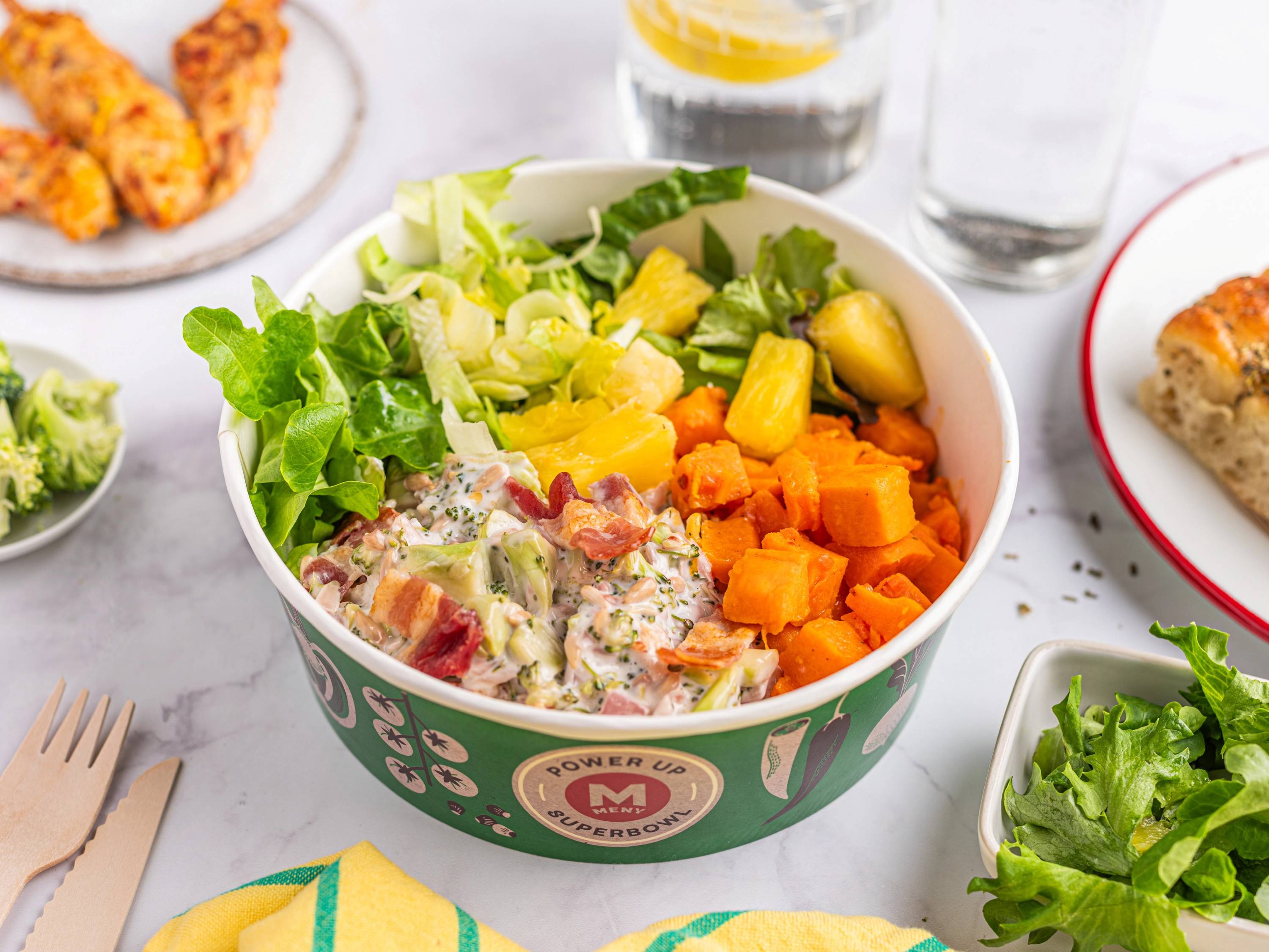 En bolle med salat og frukt, inkludert grønne blader, ananas og gulrot, på et bord med andre retter og drikkevarer rundt.